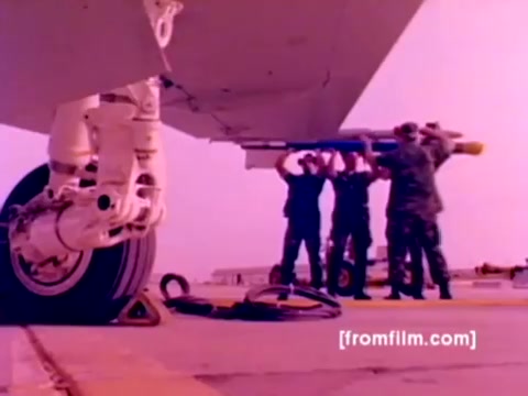 The still from the 'USMC Aviation Ordnance enlistment film' depicts a group of Marines working together to mount ordnance on an aircraft, likely an F-4 Phantom or a similar model. The scene emphasizes teamwork and the technical aspects of aircraft maintenance, showcasing military precision. The color correction enhances the visuals, bringing out the details of the aircraft's undercarriage and the ordnance being handled. It effectively highlights the roles and responsibilities involved in Marine Corps aviation ordnance operations.