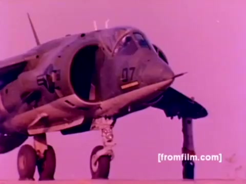 The still from the 'USMC Aviation Ordnance enlistment film' features a Harrier, prominently displaying its unique hovering capabilities. The aircraft is shown with its distinctive nose, landing gear extended, and a clear view of the weapon systems it can carry. The background likely includes a colorful, stylized depiction typical of 16mm film, possibly with a pinkish tint affecting the overall color balance. The image captures the essence of Marine Corps aviation and the focus on ordnance, highlighting the significance of this enlistment path.