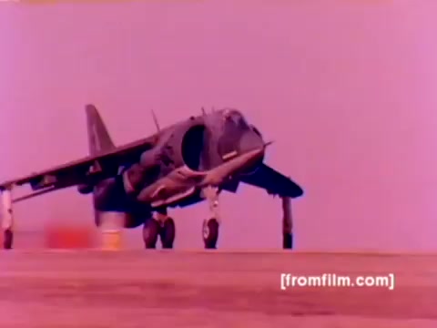 The still from the 'USMC Aviation Ordnance enlistment film' depicts a Harrier aircraft prominently in the foreground, showcasing its unique vertical takeoff capability. The color appears to be altered with a dominant pink hue, likely due to the 16mm film's age and processing. In the background, elements hint at military operations, such as ordnance being mounted, possibly missiles or bombs, emphasizing the focus on the 'Ordnance Option' for Marine Corps enlistment. The film captures the dynamic nature of Marine aviation and the critical role of ordnance in operational readiness.
