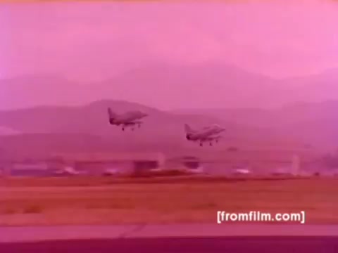 The still from the 'USMC Aviation Ordnance enlistment film' depicts two military aircraft, likely F-4 Phantoms or Harriers, taking off in the foreground. The background features a landscape with mountains, showcasing a base environment. The color is muted, likely due to the 16mm film quality, giving a vintage feel. The image highlights the dynamic action of military aviation, capturing the essence of the Ordnance Option for enlistment.   The focus on aircraft and ordnance suggests a strong emphasis on operational readiness and the importance of ordnance specialists in the Marine Corps.