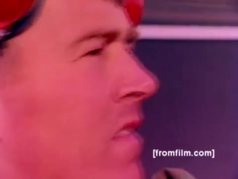 The still from the 'USMC Aviation Ordnance enlistment film' captures a close-up of a Marine, likely engaged in the process of ordnance handling. His focus suggests he is involved in preparing or inspecting military aircraft. The background hints at a dynamic environment, possibly an aircraft carrier or maintenance area.  Though silent, the imagery conveys a sense of urgency and precision associated with military operations. The color correction, when applied, would enhance the visibility of details such as the red helmet and uniform, emphasizing the operational context and the seriousness of ordnance management surrounding the aircraft like the F-4 Phantom, AH-1 Super Cobra, FA-18, and Harrier. Overall, the image reflects the crucial role of ordnance specialists in Marine Corps