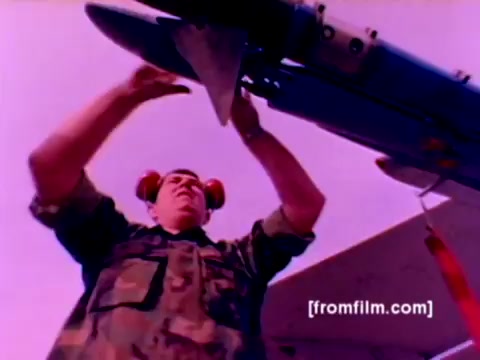 The 16mm still from the 'USMC Aviation Ordnance enlistment film' shows a Marine in camouflage fatigues and soundproof headphones engaging in the process of mounting ordnance, specifically focusing on a missile attached to an aircraft. The scene features key military aircraft, including F-4 Phantoms, AH-1 SuperCobras, an FA-18, and a Harrier, highlighting the responsibilities involved in ordnance assembly and deployment. The color may appear slightly off due to the film's age, but the image effectively captures the technical expertise required for aviation ordnance in a Marine Corps context.