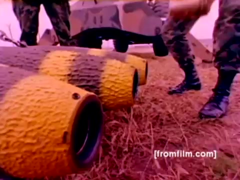 The still depicts a scene from the 'USMC Aviation Ordnance enlistment film,' showcasing essential ordnance preparations. In the foreground, we see several bombs with distinct yellow and black markings, hinting at their purpose for military use. Soldiers in camouflage uniforms are actively handling the ordnance, emphasizing the hands-on nature of the ordnance option for enlistment. The background includes military equipment, suggesting a dynamic environment focused on aviation readiness. The image has a vintage quality, reflective of the 16mm format, with a color correction applied for clarity.