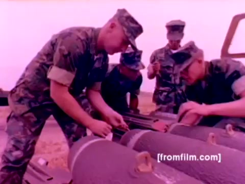 The still from the 'USMC Aviation Ordnance enlistment film' features several Marines in military fatigues working on ordnance. They are focused on mounting missiles and bombs, showcasing teamwork and technical expertise. The background includes an array of aircraft, such as F-4 Phantoms, AH-1 SuperCobras, an FA-18, and a Harrier, emphasizing the high-stakes environment of military aviation. The color correction improves the overall clarity and vibrancy, highlighting the precise nature of the ordnance work being performed.