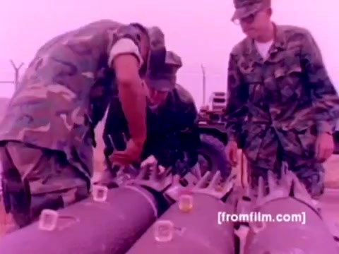 The still from the 'USMC Aviation Ordnance enlistment film' features several Marines in camouflage uniforms closely inspecting and handling ordnance. They are positioned over large bombs with multiple tails, engaged in the process of mounting missiles and bombs. The background suggests an outdoor military environment, likely near an airstrip or deployment area.  The image, derived from a 16mm negative, presents a color-corrected appearance that enhances the visual clarity of the uniforms and equipment, giving a clearer representation of the ordnance and the focus of the Marines' work. Overall, it captures a moment of technical expertise within the military aviation context.