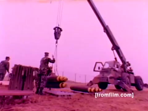 The 16mm still from the 'USMC Aviation Ordnance enlistment film' features Marine Corps personnel engaged in ordnance operations. In the scene, a crane is used to lift munitions—specifically missiles and bombs—while military members supervise and assist. The background suggests an outdoor military environment.   The overall color appears to be somewhat washed out, giving it a vintage look typical of early film formats. The focus on ordnance preparation highlights the technical aspects of the Ordnance Option for enlistment.