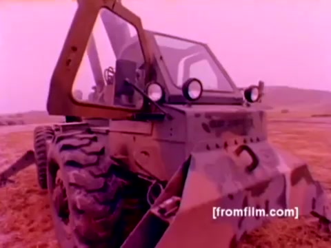 The still from the 'USMC Aviation Ordnance enlistment film' shows a military vehicle, possibly used for mounting ordnance, in a military camouflage pattern. The vehicle has large tires and a raised, protective cabin with multiple lights. The background suggests an open landscape, likely used for operational training or demonstrations. The image has a color tint that could be a result of aging in the 16mm film, creating a pinkish hue. Overall, it reflects the military's emphasis on aviation ordnance capabilities within Marine Corps enlistment options.