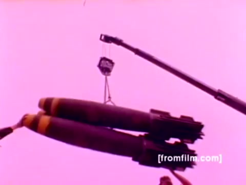 The still from the USMC Aviation Ordnance enlistment film captures a moment of ordnance loading, showcasing two large bombs being lifted by a crane. The background features a distinct pink hue, likely due to the 16mm negative's color correction. This moment highlights the operational side of Marine aviation, emphasizing the ordnance capability associated with aircraft like the F-4 Phantom, AH-1 Super Cobra, FA-18, and Harrier, which are depicted throughout the film. The focus on ordnance mounting underscores the importance of preparedness and operational efficiency in Marine aviation.