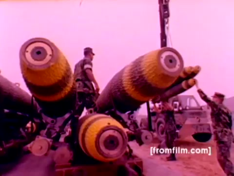 The still captures a scene from the 'USMC Aviation Ordnance enlistment film,' showcasing Marines involved in the ordnance support role. Featured are large ordnance pieces, likely bombs or missile systems, with distinct yellow and black markings. Marines are actively engaged in mounting the ordnance, demonstrating teamwork and precision. The background suggests a military environment, possibly a deployment area, with military vehicles present. The color correction improves the overall visibility while retaining the archival quality of the 16mm film.