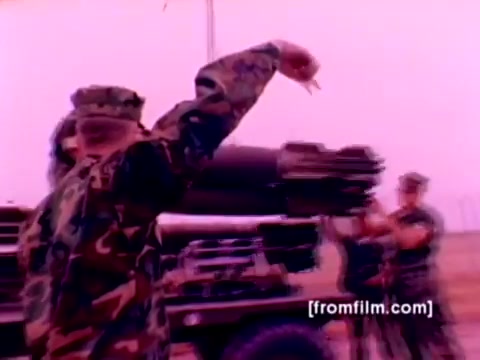The still from the 'USMC Aviation Ordnance enlistment film' depicts a military scenario focused on ordnance handling. It features a Marine in camouflage uniform gesturing, likely directing others as they prepare to mount weapons. In the background, fellow Marines are present, working on the logistics of loading missiles and bombs onto aircraft. The aircraft shown include F-4 Phantoms, AH-1 SuperCobras, an FA-18, and a Harrier, emphasizing the ordnance options available for enlistment in the Marine Corps. The overall color correction would enhance details and clarity, making the operational context more visually engaging.