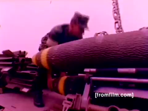The 16mm still from the 'USMC Aviation Ordnance enlistment film' features a Marine in uniform handling ordnance. He is seen mounting large bombs, possibly preparing them for attachment to aircraft. Various military aircraft such as F-4 Phantoms, AH-1 SuperCobras, FA-18s, and Harriers are present in the film's context. The image has a pinkish tint, likely due to the film's original color processing. A re-render with better color correction would enhance the visual clarity and provide a more accurate representation of the scene.