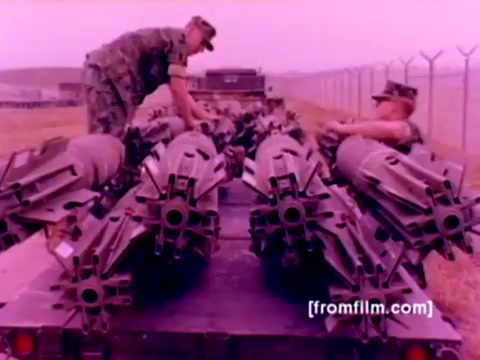 The still from the 'USMC Aviation Ordnance enlistment film' captures a scene featuring U.S. Marine Corps personnel working on a truck bed loaded with missiles. The image shows two Marines in fatigue uniforms inspecting and preparing the ordnance, highlighting the hands-on nature of aviation ordnance training. The background includes a fenced area typical of military installations, with a distant view of aircraft like F-4 Phantoms, AH-1 SuperCobras, FA-18s, and Harriers suggested in the context of the film. The color appears to have a pink tint, likely due to the film’s age and the 16mm format, suggesting a need for better color correction for clarity and vibrancy.