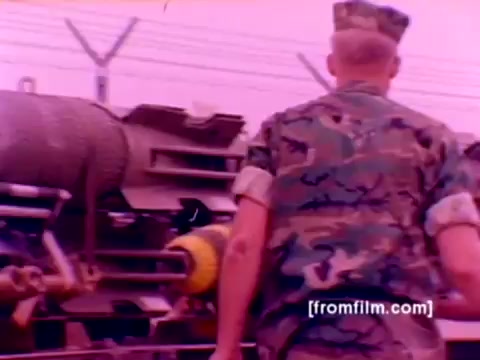 The still from the 'USMC Aviation Ordnance enlistment film' features a Marine in camouflage uniform walking towards a military vehicle, which appears to be loaded with ordnance equipment. The background may include aircraft such as F-4 Phantoms, AH-1 SuperCobras, an FA-18, and a Harrier. The image captures the essence of military operations related to ordnance mounting, with a focus on missiles and bombs. The color correction enhances the visual clarity, reflecting the aircraft's significance in Marine Corps aviation.