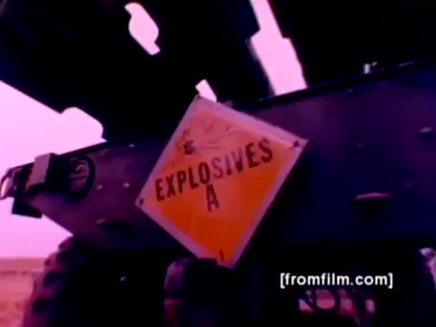 The still from the 'USMC Aviation Ordnance enlistment film' captures a vivid moment focusing on ordnance operations within the Marine Corps. The scene features a sign clearly marked 'EXPLOSIVES A,' likely indicating the presence of munitions being handled.   In the context of the film, it highlights the technical environment involving various aircraft such as F-4 Phantoms, AH-1 SuperCobras, FA-18s, and Harriers, as they undergo ordnance mounting with missiles and bombs. The image is tinted with a washed-out pink hue, typical of 16mm negatives, which could benefit from improved color correction for clarity and realism. Overall, it conveys a serious and operational atmosphere in military aviation.