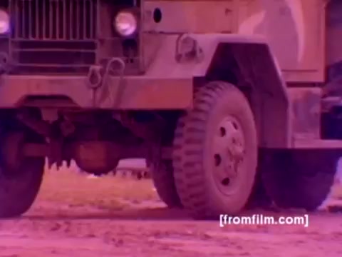 The still from the 'USMC Aviation Ordnance enlistment film' features military vehicles, likely in a training or operational environment. The focus appears to be on a military truck, illustrating its rugged design with large, sturdy tires suited for diverse terrains. The color is less vibrant, typical of 16mm film, but with color correction, it would present a more realistic rendering, enhancing the military greens and earth tones. The context suggests this scene serves to highlight the logistical aspects of ordnance handling, showcasing the Marine Corps' focus on aviation and ordnance readiness.
