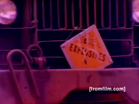 The still from the 'USMC Aviation Ordnance enlistment film' features a close-up of a military vehicle's grill displaying a bright orange sign labeled 'EXPLOSIVES.' The background showcases a vibrant but somewhat muted color palette, typical of a 16mm film negative. The image suggests a focus on safety and ordinance handling, with the intention to highlight the responsibilities associated with the Ordnance Option in Marine Corps enlistment.