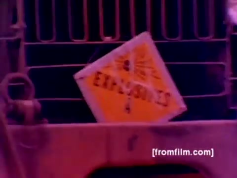 The still portrays a military vehicle's rear, prominently displaying a cautionary 'EXPLOSIVES' sign. The image has a vintage 16mm feel, with color correction needed to enhance clarity. The background suggests a military environment, potentially connected to the handling of ordnance, consistent with the film's focus on the Marine Corps' Ordnance Option. The presence of this sign emphasizes the critical nature of safety in military operations involving weapons and explosives.