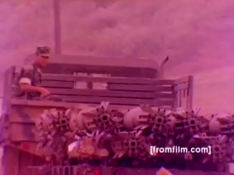 The still from the 'USMC Aviation Ordnance enlistment film' likely depicts a Marine in military attire, overseeing the mounting of ordnance such as missiles and bombs onto aircraft. Visible are various military aircraft like F-4 Phantoms, AH-1 SuperCobras, an FA-18, and a Harrier, indicating a focus on aviation ordnance. The scene is characterized by a somewhat faded color palette, typical of a 16mm negative, which could benefit from improved color correction for clarity and detail.