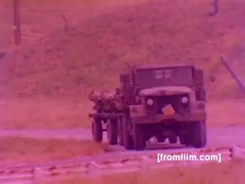 The still from the 'USMC Aviation Ordnance enlistment film' captures a military scene featuring a truck transporting ordnance along a road. The background showcases a subtle landscape with a mix of grass and terrain, typical of military training grounds. The color is slightly off, with a pinkish tint, likely due to the 16mm negative’s aging, impacting the overall clarity and vibrancy of the imagery. Although silent, the visuals convey the action-oriented nature of the Marine Corps and highlight the significance of ordnance operations, as seen with the diverse array of aircraft like the F-4 Phantom, AH-1 SuperCobra, FA-18, and Harrier being discussed in the context of enlistment options.
