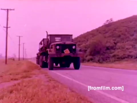 The still from the 'USMC Aviation Ordnance enlistment film' depicts a military truck on a roadway, set against a backdrop of rolling hills. The film likely features elements related to Marine Corps aviation ordnance, showcasing aircraft such as F-4 Phantoms, AH-1 SuperCobras, FA-18s, and Harriers. The focus appears to be on ordnance being mounted, including missiles and bombs. The color correction suggests a vintage aesthetic typical of a 16mm negative, but a more vibrant hue would enhance the visual experience. Overall, the image evokes a sense of military readiness and equipment handling.