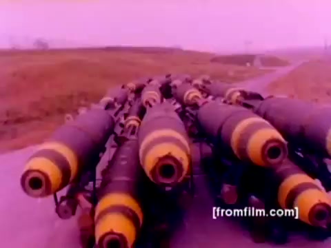 The 16mm still from the 'USMC Aviation Ordnance enlistment film' shows a lineup of ordnance, specifically missiles and bombs, prominently featuring those with distinct yellow markings. The composition emphasizes the military hardware, suggesting a focus on preparation and functionality in aviation operations. The colors appear muted due to the aging of the film, lacking the vibrancy typical of modern visuals. Overall, the image encapsulates military readiness within the context of Marine Corps aviation.