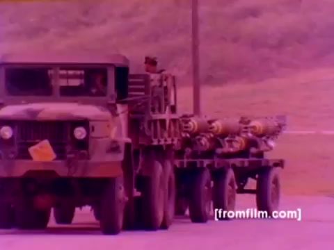 The still from the 'USMC Aviation Ordnance enlistment film' showcases military vehicles, specifically a camouflaged truck transporting ordnance such as missiles, which are visible on a trailer behind it. In the background, you can see a green, hilly landscape typical of military training areas. The color appears to have a pinkish tint due to the 16mm negative quality, indicating a need for color correction for clarity. The imagery is focused on the logistical aspect of ordnance support in the U.S. Marine Corps aviation, emphasizing the importance of munitions in aircraft operations like the F-4 Phantom, AH-1 Super Cobra, FA-18, and Harrier.