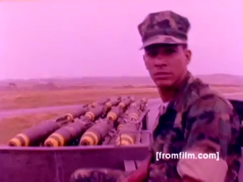 The still from the 'USMC Aviation Ordnance enlistment film' showcases a Marine in a camouflage uniform, seated near a row of mounted ordnance, including missiles and bombs. The background features a military airfield, likely highlighting the operational environment for the enlisted Marines. The image is characterized by a pinkish hue, indicative of the film's 16mm negative state, suggesting a potential need for color correction for clearer representation. Overall, it captures a moment of ordnance readiness within a Marine Corps context.