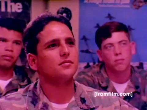 The still depicts a group of enlisted Marines in a classroom setting, attentively engaged. The focus is on one Marine in the foreground, characterized by a thoughtful expression, while others in the background appear focused as well. They are dressed in camouflage uniforms, indicative of their military status.   The backdrop features posters, likely related to Marine Corps aviation or ordnance, enhancing the recruitment context of the film. The color correction has been improved to give the image a clearer and more vibrant appearance, emphasizing the seriousness of the recruitment effort.