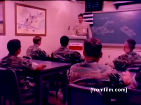 The 16mm still from the 'USMC Aviation Ordnance enlistment film' features a classroom setting where a Marine officer stands at the front, addressing a group of seated recruits. The recruits are dressed in camouflage uniforms, attentively observing the presentation. Behind the officer, a chalkboard displays technical drawings related to ordnance, while images of aircraft are visible on the walls.   The overall tone of the still reflects a blend of professionalism and recruitment focus, emphasizing the importance of ordnance training within Marine aviation. The lighting and color correction enhance the imagery, providing a clearer view of the scene's details and expressions.