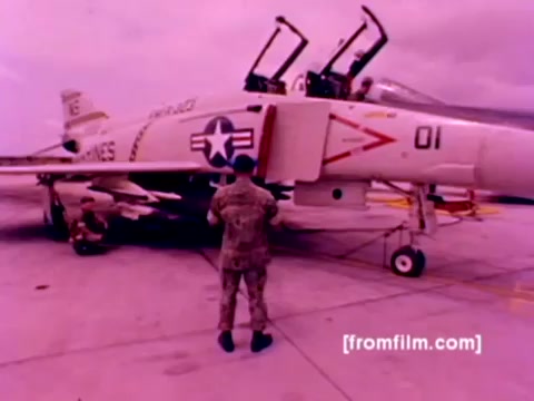 The 16mm still from the 'USMC Aviation Ordnance enlistment film' showcases a Marine Corps scene featuring an F-4 Phantom aircraft. A Marine, dressed in uniform, is seen interacting with the aircraft on the tarmac. The jet is detailed with military markings, including a star insignia and a unique tail number.   In this context, the film emphasizes the 'Ordnance Option' for enlistment, highlighting the mounting of missiles and bombs on various military aircraft, including F-4s, AH-1 SuperCobras, FA-18s, and Harriers. The color correction enhances the aircraft's details and gives the image a more realistic appearance. Overall, it captures the technical aspects of Marine aviation and ord