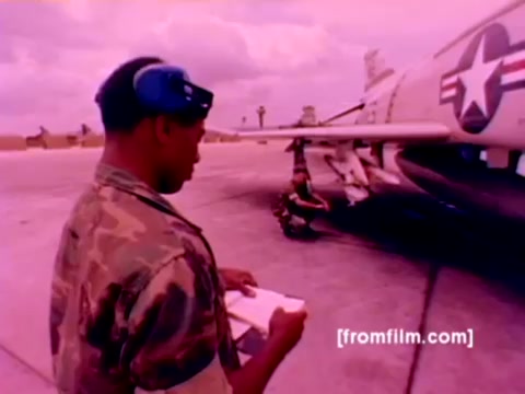 This 16mm still from the 'USMC Aviation Ordnance enlistment film' depicts a military setting with an F-4 Phantom in the background. In the foreground, a Marine, wearing camouflage and a headset, is reviewing a clipboard. Another Marine is visible, engaged in the process of mounting ordnance, such as missiles and bombs, on the aircraft. The image has a strong pink hue, typical of older film stock, making color correction necessary for clarity and realism. The scene reflects the operational aspects of Marine Corps aviation ordnance work, emphasizing the technical and teamwork elements involved in military aviation.