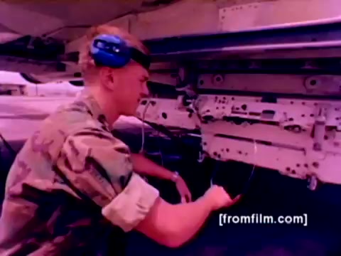 The still from the 'USMC Aviation Ordnance enlistment film' depicts a U.S. Marine in uniform, outfitted with a headset, as he works on mounting ordnance. The setting includes aircraft like F-4 Phantoms, AH-1 SuperCobras, an FA-18, and a Harrier, which are all part of the Marine Corps fleet. This moment captures the technician involved in intricate procedures surrounding missile and bomb installation, emphasizing the technical aspects of aviation ordnance roles. The image is rendered from a 16mm negative, with color correction enhancing its visual clarity and vibrancy.