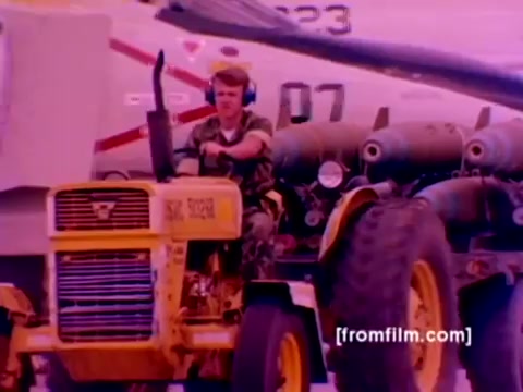 The still from the 'USMC Aviation Ordnance enlistment film' features a scene on a military airfield with a focus on aviation ordnance operations. In the foreground, a Marine is operating a yellow tractor, likely involved in transporting ordnance. The background showcases military aircraft, including an F-4 Phantom, AH-1 SuperCobra, FA-18, and Harrier.   Ordnance, such as missiles and bombs, is visible, indicating the film's emphasis on the precision and responsibilities of ordnance personnel. The image has a somewhat muted color palette typical of old 16mm film, reflecting the era’s film quality. Overall, it conveys the critical role of aviation ordnance within the Marine Corps.