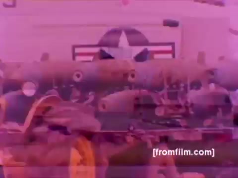 The 16mm still from the 'USMC Aviation Ordnance enlistment film' showcases a dynamic scene involving military aircraft, specifically F-4 Phantoms, AH-1 SuperCobras, an FA-18, and a Harrier. The focus is on the ordnance being mounted, highlighting the meticulous process of attaching missiles and bombs to these aircraft. The color rendering appears faded or incorrectly cast, likely due to the age of the negative, giving the image a pinkish hue. Overall, it captures the technical and operational aspects of Marine Corps aviation ordnance.