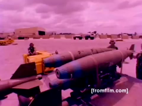 The still from the 'USMC Aviation Ordnance enlistment film' showcases a military scene with ordnance, specifically missiles and bombs, being prepared for aircraft. F-4 Phantoms, AH-1 SuperCobras, an FA-18, and a Harrier are part of the context, highlighting the Marine Corps' aviation capabilities. The image likely has a warm, heavily tinted color cast typical of old 16mm film, indicating the need for better color correction to enhance the details and clarity of the equipment and personnel involved in the ordnance handling process.