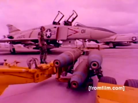 The still from the 'USMC Aviation Ordnance enlistment film' features a military setting with various aircraft in the background, including F-4 Phantoms and an FA-18. In the foreground, a service member operates machinery used for mounting ordnance, such as missiles and bombs. The colors in the image appear oversaturated, creating a pinkish hue, likely due to the original 16mm film quality. A clearer color correction could enhance the visibility of details, showcasing the military equipment and personnel more effectively. The scene emphasizes the technical aspects of Marine Corps aviation ordnance operations.