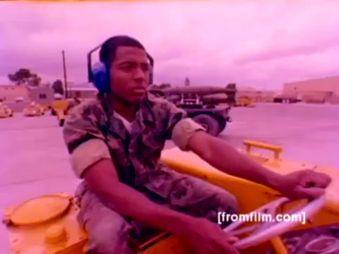 The still features a US Marine in camouflage fatigues operating a yellow ground vehicle, presumably involved in ordnance handling. He wears blue headphones, indicating the noisy environment typical of a military base. The background includes other vehicles and equipment, hinting at an active ordnance loading area. The color correction reveals a washed-out, surreal palette typical of older 16mm film, enhancing the historical feel of the Marine Corps enlistment context. Overall, the scene captures the precision and responsibility associated with ordnance operations.