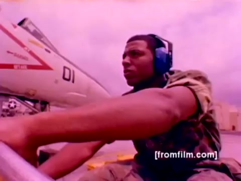 The still from the 'USMC Aviation Ordnance enlistment film' depicts a U.S. Marine in military fatigues, wearing headphones and focused on his task. He is positioned in front of an F-4 Phantom, with other military aircraft likely featuring in the film, including AH-1 SuperCobras, FA-18s, and Harriers. The scene shows the mounting of ordnance, specifically missiles and bombs, signifying the technical role of ordnance specialists in aviation. The image captures the intensity and professionalism associated with Marine Corps aviation operations, presented with an outdated color palette typical of 16mm film.