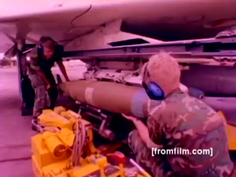 The still from the 'USMC Aviation Ordnance enlistment film' showcases two Marines working on the ground, focused on mounting ordnance onto an aircraft. One Marine is handling a large missile, positioned on a yellow support structure, while another assists nearby, wearing protective headgear. The background features part of the aircraft, likely an F-4 Phantom or similar, indicating an operational environment. The 16mm negative gives the image a vintage feel, and despite the lack of sound, the scene conveys the critical role of ordnance maintenance in Marine aviation. Enhanced color correction could reveal more details and vibrancy in the military uniforms and ordnance involved.
