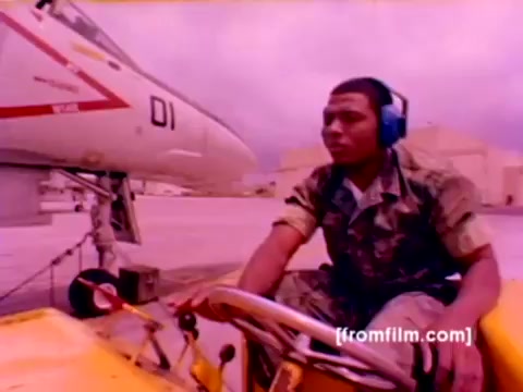 The still from the 'USMC Aviation Ordnance enlistment film' captures a Marine in a military uniform operating a yellow ground vehicle near an F-4 Phantom aircraft. The background features military aviation equipment and the aircraft itself, which is partially visible with a prominent red and white design. The Marine wears headphones, indicating focus on his task. The image is part of a silent film showcasing the Ordnance Option, emphasizing the technical and operational aspects of ordnance management, including the handling of missiles and bombs. The overall color appears washed out, typical of 16mm negatives, which would benefit from better correction to enhance visual clarity.