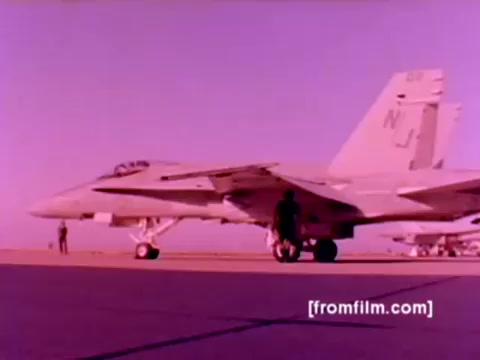 The still from the 'USMC Aviation Ordnance enlistment film' features prominent military aircraft including F-4 Phantoms, AH-1 SuperCobras, an FA-18, and a Harrier. In the scene, ordnance personnel are seen mounting missiles and bombs onto the aircraft, highlighting the ordnance operation within the Marine Corps. The image has been pulled from a 16mm negative, presenting the aircraft in vivid colors. The overall atmosphere conveys the precision and technical expertise required in military ordnance operations.