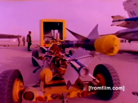 The still from the 'USMC Aviation Ordnance enlistment film' features military aircraft, including F-4 Phantoms and an AH-1 SuperCobra, in a vivid outdoor setting. In the foreground, an advanced ordnance cart showcases missiles and bomb mounts, emphasizing the technical aspects of ordnance handling. A Marine in uniform stands nearby, indicating the operational context. The color correction would enhance the distinct features of the aircraft and ordnance, emphasizing details and clarity in this marine aviation setting.