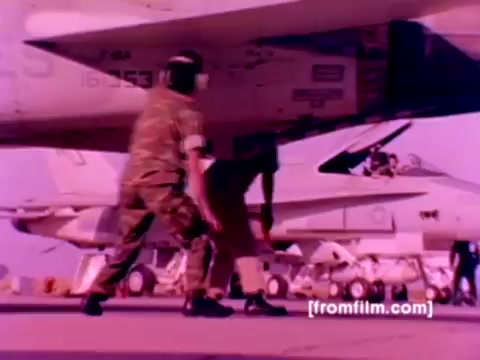 This still from the 'USMC Aviation Ordnance enlistment film' features U.S. Marines working on various aircraft, including F-4 Phantoms, AH-1 SuperCobras, FA-18s, and Harriers. The scene highlights the ordnance being mounted, such as missiles and bombs, showcasing the technical skills involved in aircraft maintenance and armament. The color appears saturated due to the 16mm negative, requiring color correction for a more accurate representation. The image captures a dynamic and professional environment typical of military aviation operations.
