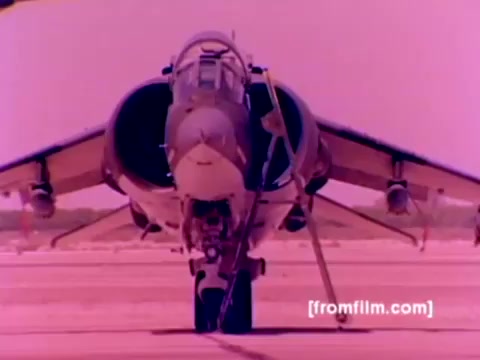 The still from the 'USMC Aviation Ordnance enlistment film' showcases a Harrier aircraft prominently, with its iconic vertical takeoff design. The scene is likely set on an airstrip, emphasizing the military's focus on ordnance, as indicated by the presence of missiles and bombs being mounted on various aircraft, including F-4 Phantoms, AH-1 SuperCobras, and FA-18s. The colors appear overly saturated, suggesting the film's age and the need for better color correction to reflect the aircraft's true appearance. Overall, the still captures the essence of Marine Corps aviation and ordnance operations.