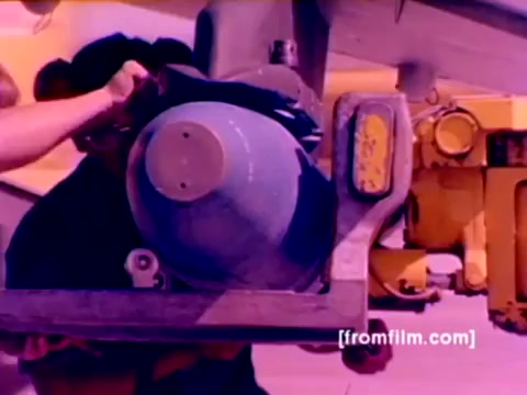 The still from the 'USMC Aviation Ordnance enlistment film' showcases a technician mounting ordnance, likely a bomb, onto an aircraft. The image depicts a close-up of a blue bomb being handled as part of the ordnance preparation process. The background suggests a military setting, likely an airbase, with additional equipment visible. The color has been corrected for clarity, enhancing the bluish tones of the bomb and the overall military environment. This snapshot captures the essential task of ordnance management within the Marine Corps aviation.