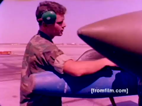 The still features a Marine in camouflage uniform, working on an aircraft. He is wearing green headphones and is focused on the blue section of the aircraft, likely preparing to mount ordnance. The background hints at an airfield setting, indicative of a military environment. The colors suggest a likely enhancement to bring out more vibrancy from the original 16mm film, providing a clearer view of the aircraft and the working conditions. Overall, it captures the essence of Marine Corps aviation and ordnance operations.