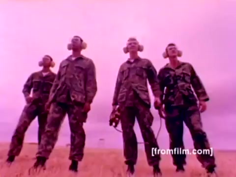 The still from the 'USMC Aviation Ordnance enlistment film' features four Marines standing in military uniforms and headsets, positioned against a surreal, muted pink sky, likely highlighting an emphasis on their readiness and focus. The context involves showcasing the Ordnance Option for enlistment, with additional scenes in the film displaying various aircraft, including F-4 Phantoms, AH-1 SuperCobras, an FA-18, and a Harrier, alongside the mounting of missiles and bombs. The image captures a moment of camaraderie and determination among the Marines involved in ordnance operations.