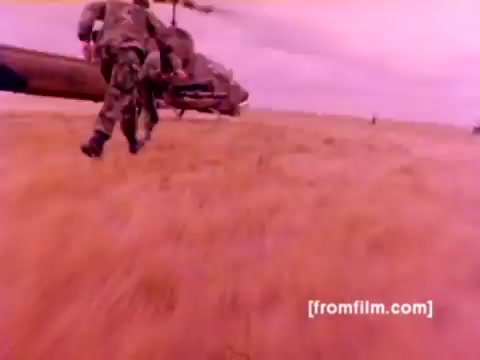 The 16mm still from the 'USMC Aviation Ordnance enlistment film' depicts Marine Corps personnel in camouflage gear rushing toward an AH-1 SuperCobra helicopter in a field. The scene conveys urgency and action, highlighting the operation of aircraft like the F-4 Phantom and FA-18, as well as the Harrier. The focus is on ordnance being mounted, showcasing missiles and bombs. The color correction from the original negative emphasizes a warmer, more vivid palette, bringing out details in the uniforms and environment. The overall impression is one of military readiness and the dynamic role of ordnance in aviation.