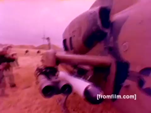 The 16mm still from the 'USMC Aviation Ordnance enlistment film' depicts a close-up view of an AH-1 SuperCobra helicopter, showcasing ordnance being mounted, including missiles. The background hints at military personnel working with the aircraft, emphasizing the operational aspect of Marine Corps aviation. The color appears heavily tinted, likely due to the aging of the film, giving it a purple hue. Overall, the still effectively captures the technical and collaborative environment of aviation ordnance within the Marine Corps.