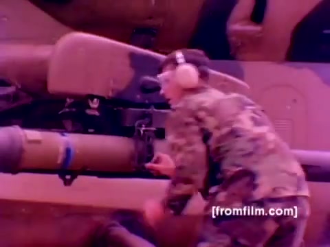 The still from the 'USMC Aviation Ordnance enlistment film' features a Marine in uniform, working on the ordnance of a hovering AH-1 Super Cobra helicopter. He is shown mounting a missile, wearing protective earphones and focused on the task. The vivid colors highlight the operational environment, showcasing the high-tech military equipment typical of the Marine Corps. The background suggests a busy airfield or maintenance area, emphasizing the dynamic nature of ordnance work.